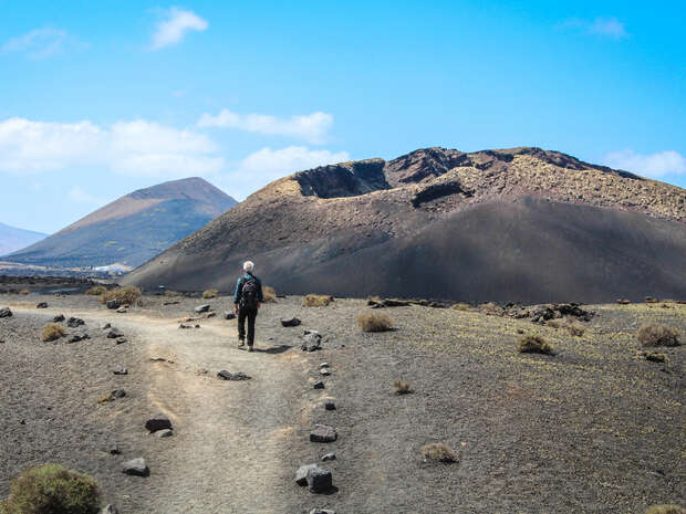 A person with a backpack walks on a rocky path through a barren, volcanic landscape, surrounded by dry, dark hills under a clear blue sky.